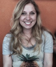 Lächelnde Frau mit langen, gewellten Haaren trägt ein T-Shirt mit Ananas-Design, steht vor orangefarbener Wand, repräsentiert kreative Handwerkskunst und Kultur.