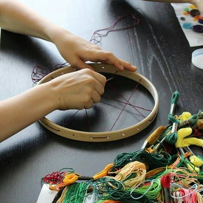 Personen arbeiten an einem Holzrahmen für einen Traumfänger, umgeben von bunten Fäden und Perlen, kreative Handarbeit für individuelle Traumfänger.