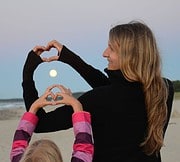 Frau und Kind formen Herzzeichen mit den Händen, während die Sonne am Himmel untergeht, symbolisiert Liebe und Verbundenheit bei HANDMADE Kultur.