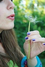 Frau hält eine Pusteblume, Symbol für Natur und Handwerkskunst, mit grüner Natur im Hintergrund, betont die Verbindung zu nachhaltigem und kreativem Handwerk.