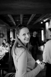Fröhliche Frau hält Smartphone in der Hand, in einem gemütlichen, handwerklich gestalteten Café, umgeben von anderen Gästen, im schwarzen und weißen Stil.