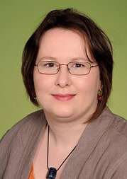 Porträt einer Frau mit Brille und kurzen braunen Haaren vor grünem Hintergrund, freundliches Lächeln, professionell und einladend, ideal für Handmade- und Kulturthemen.
