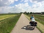 Frau fährt mit dem Fahrrad auf einer geraden, asphaltierte Strecke durch eine grüne Landschaft mit Feldern und Bäumen, bei sonnigem Himmel.