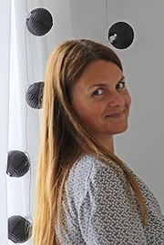 Frau mit langen braunen Haaren, die neben einer Wand mit schwarzen, runden Dekorationen steht, symbolisiert kreative Handwerkskunst und DIY-Projekte bei HANDMADE Kultur.