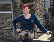 Frau mit rotem Haar, bunter Kleid und blauer Bluse posiert vor einer Backsteinmauer bei Handmade Kultur, umgeben von Natur und urbanem Ambiente.