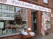 Geschäft für Patchwork, Quilting, Stoffe und Handarbeitszubehör mit Schaufenster und Sitzbank vor dem Laden.