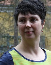 Frau mit kurzen dunklen Haaren trägt ein bunt gestreiftes Oberteil und schaut nach rechts, im Hintergrund Bäume und eine Stadtumgebung.