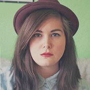 Porträt einer jungen Frau mit braunem Haar, rotem Lippenstift und Hut, vor grünem Hintergrund, im Stil von Handmade Kultur, Fokus auf kreative und handgemachte Mode.