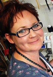 Frau mit roten Haaren und Brille, die an einem Nähprojekt arbeitet, umgeben von Nähmaschinen und Stoffen, bei Handmade Kultur.