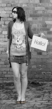 Frau mit dunklen Haaren, Sonnenbrille, T-Shirt mit Hasenmotiv, kurzer Rock, Cardigan, hält ein Schild mit 'Hallo!', vor einer Ziegelmauer, bei Handmade Kultur.