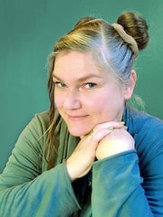 Frau mit kreativ geflochtenem Haar, die ihre Hände unter dem Kinn verschränkt und in die Kamera lächelt, symbolisiert handgemachte Kunst und kreative Handwerkskunst.