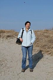 Frau mit Rucksack beim Spaziergang an der Küste, trägt eine weiße Jacke und Jeans, umgeben von Sanddünen und blauer Himmel, entspannte Atmosphäre, Natur, Freizeit, Urlaub.
