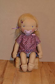 Handgemachte Stoffpuppe mit rosa Kleid, rosa Haarband und Schnuller, sitzend auf einem kleinen Korb, ideal für Handmade- und Bastelkunst.