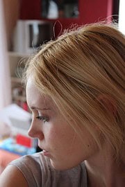 Nahaufnahme einer jungen Frau mit roten Haaren, die nachdenklich schaut, im Hintergrund ein wohnliches Umfeld mit Bücherregal und Dekorationen.