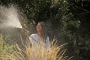 Frau beim Gießen im Garten, umgeben von Pflanzen und Büschen, mit Wasser sprühend für eine gepflegte Gartenlandschaft.