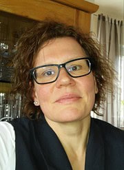 Porträt einer Frau mit Brille, kurze lockige Haare, lächelnd, in einem gemütlichen Raum mit Holzregal und Vorhängen, Fokus auf Gesicht und Ausdruck.