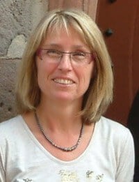 Eine Frau mit Brille und Schulterlänge blondem Haar, lächelnd, trägt ein weißes Oberteil und eine Halskette, steht vor einer Steinmauer, symbolisiert kreative Handwerkskunst.