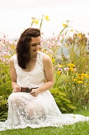 Frau in weißem Kleid sitzt im Garten mit bunten Blumen, umgeben von Natur und Blüten, kreative Handwerkskunst, nachhaltige Mode, Naturverbundenheit, entspannte Atmosphäre.