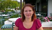 Frau mit roten Haaren in einem pinken Top sitzt in einem Gartenrestaurant mit Blumen und Tischen, die für handgemachte Kulturveranstaltungen vorbereitet sind.