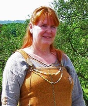 Frau mit rotem Haar trägt traditionelle handgemachte Kleidung und Schmuck, umgeben von grüner Natur, symbolisiert Handwerkskunst und kulturelle Traditionen.