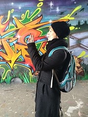 Junge Frau mit Rucksack betrachtet Graffiti-Kunst an einer Wand, trägt schwarze Mütze und Mantel, urbanes Umfeld, kreative Street Art Szene, Lifestyle und Kultur.
