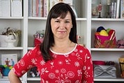 Frau in rotem Handarbeits-Shirt vor einem Regal mit bunten Wolle- und Bastelmaterialien, passend für kreative DIY-Projekte und Handarbeit.