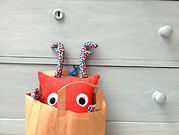 Kuscheliges Stofftier mit großen Augen und bunten Antennen, in einer braunen Papiertüte, als handgemachtes Kunsthandwerk, ideal für Dekoration oder Geschenk.