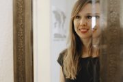 Frau mit langem blondem Haar und rotem Lippenstift schaut in einen Spiegel, vermittelt handgemachte Kultur und kreative Handwerkskunst.