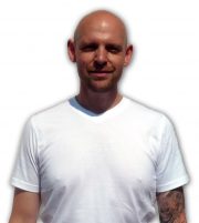 Mann mit Tattoos auf dem linken Arm, trägt ein weißes T-Shirt, lächelt leicht, steht vor einem weißen Hintergrund, Fokus auf Person, für Handmade Kultur SEO optimiert.