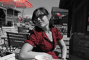 Frau mit Brille und rotem Karohemd sitzt in einem Outdoor-Café, umgeben von Holzstühlen und Tischen, mit einem roten Sonnenschirm im Hintergrund, bei Handmade Kultur.
