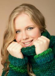 Junge Frau trägt handgestrickte grüne und blaue Pullover, lächelt freundlich, zeigt die Textur der handgemachten Strickwaren, betont die kreative Handarbeit bei Handmade Kultur.