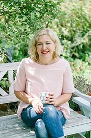 Frau mit blonden, lockigen Haaren sitzt auf einer Bank im Garten, hält eine Tasse mit Punkten, umgeben von grünen Pflanzen, für Handmade Kultur SEO optimiert.