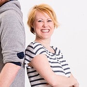 Lächelnde Frau mit kurzen roten Haaren in gestreiftem T-Shirt, neben einem Mann in grauem Pullover, beide zeigen Freude und Verbundenheit, symbolisieren Handmade Kultur.