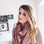 Junge Frau mit langem, blondem Haar trägt einen pinkfarbenen Schal und ein Musteroberteil, umgeben von Kunstwerken an der Wand, symbolisiert kreative Handwerkskunst und Mode.