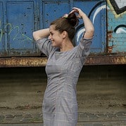 Frau mit hochgestecktem Haar in einem karierten Kleid, die lächelnd ihre Haare bindet, vor einer graffiti-beschmierten Wand und einem alten Zugwagen.