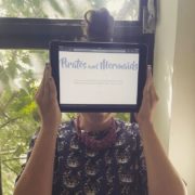 Person hält ein Tablet vor das Gesicht, auf dem die Webseite 'Pirates and Mermaids' zu sehen ist, mit einem Fenster im Hintergrund, das Natur zeigt.