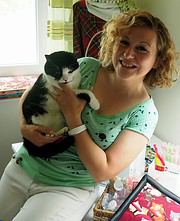 Frau mit lockigem Haar hält eine schwarz-weiße Katze in den Händen, in einem hellen Raum mit bunten Dekorationen und Bastelmaterialien im Hintergrund.
