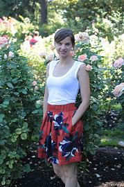 Frau in einem weißen Top und rotem Rock mit Blumenmuster, umgeben von blühenden Rosen im Garten, bei sonnigem Wetter, entspannt und glücklich.