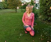 Frau in bunter, handgemachter Strickjacke und bunten Leggings kniet im Garten, umgeben von blühenden Rosen und grüner Natur, bei Handmade Kultur.