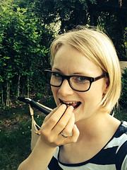 Frau mit Brille und kurzen blonden Haaren, die im Garten lächelnd eine Hand vor den Mund hält, umgeben von grünen Pflanzen und Bäumen.