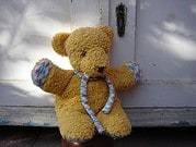 Handgefertigter Plüschbär mit buntem Schal, der vor einer weißen Tür sitzt, ideal für Handmade- und Kuschelkunst-SEO.