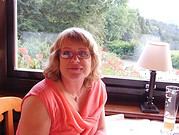 Frau mit Brille und lockigem Haar sitzt an einem Tisch vor einem Fenster mit Blick auf einen Garten, umgeben von handgemachten Kunstwerken und kreativen Materialien.