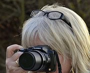 Fotografin mit weißem Haar, Brille auf Kopf, hält Kamera beim Fotografieren, Fokus auf professionelles Equipment, kreative Arbeit im Freien.