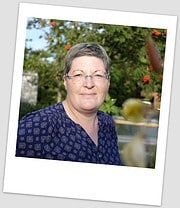 Porträt einer Frau mit kurzen grauen Haaren und Brille, trägt eine blaue Bluse mit Muster, im Garten mit grünen Bäumen und roten Blumen im Hintergrund.