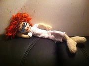 Handgefertigte Stoffpuppe mit rotem Haar, Brille und gestricktem Kleid, liegend auf einer schwarzen Couch, kreative Handarbeit, Kunsthandwerk, Dekoration.