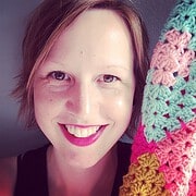 Fröhliche Frau mit kurzen Haaren und rotem Lippenstift, hält eine bunte gehäkelte Decke, zeigt kreative Handarbeit und Kunsthandwerk bei Handmade Kultur.