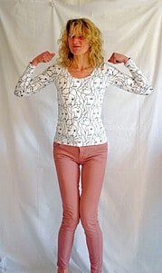 Frau mit lockigem blondem Haar trägt ein langärmeliges Shirt mit abstrakten Gesichtern und rosa Hosen, präsentiert ihre Muskeln in einer selbstbewussten Pose.