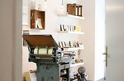 Alte Druckmaschine in einer kreativen Werkstatt, umgeben von Regalen mit Büchern und Dekorationsgegenständen, im Fokus auf handwerkliche Druckkunst und kreative Gestaltung.