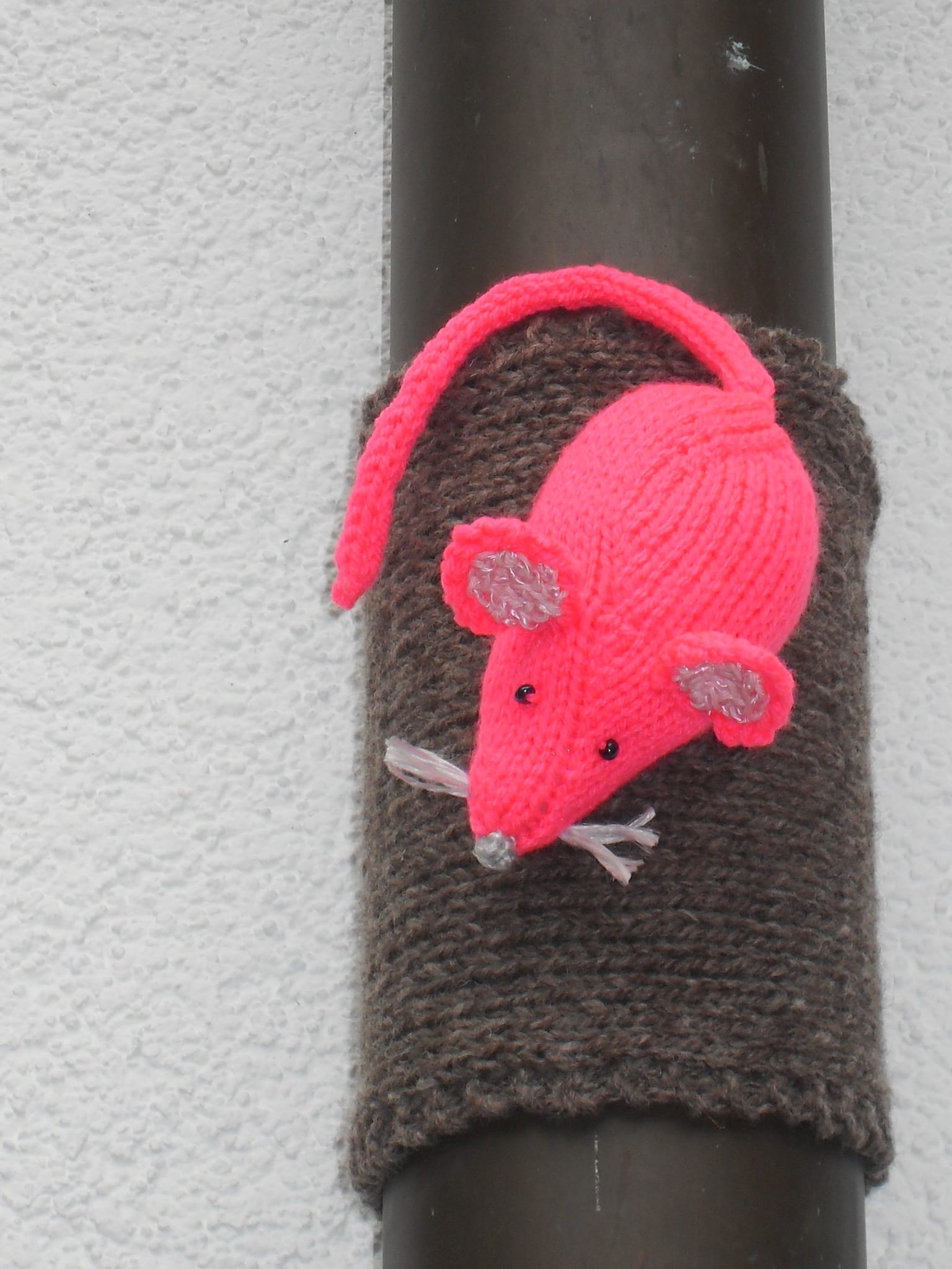 Handgefertigte, gestrickte Maus aus Wolle in Pink und Grau, dekorativ an einem schwarzen Rohr befestigt, ideal für kreative Handwerkskunst und Dekorationen.