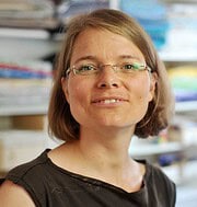 Frau mit Brille und freundlichem Lächeln in einem Handwerksladen, umgeben von bunten Stoffen und Bastelmaterialien, die handgemachte Kunst und Kreativität repräsentieren.